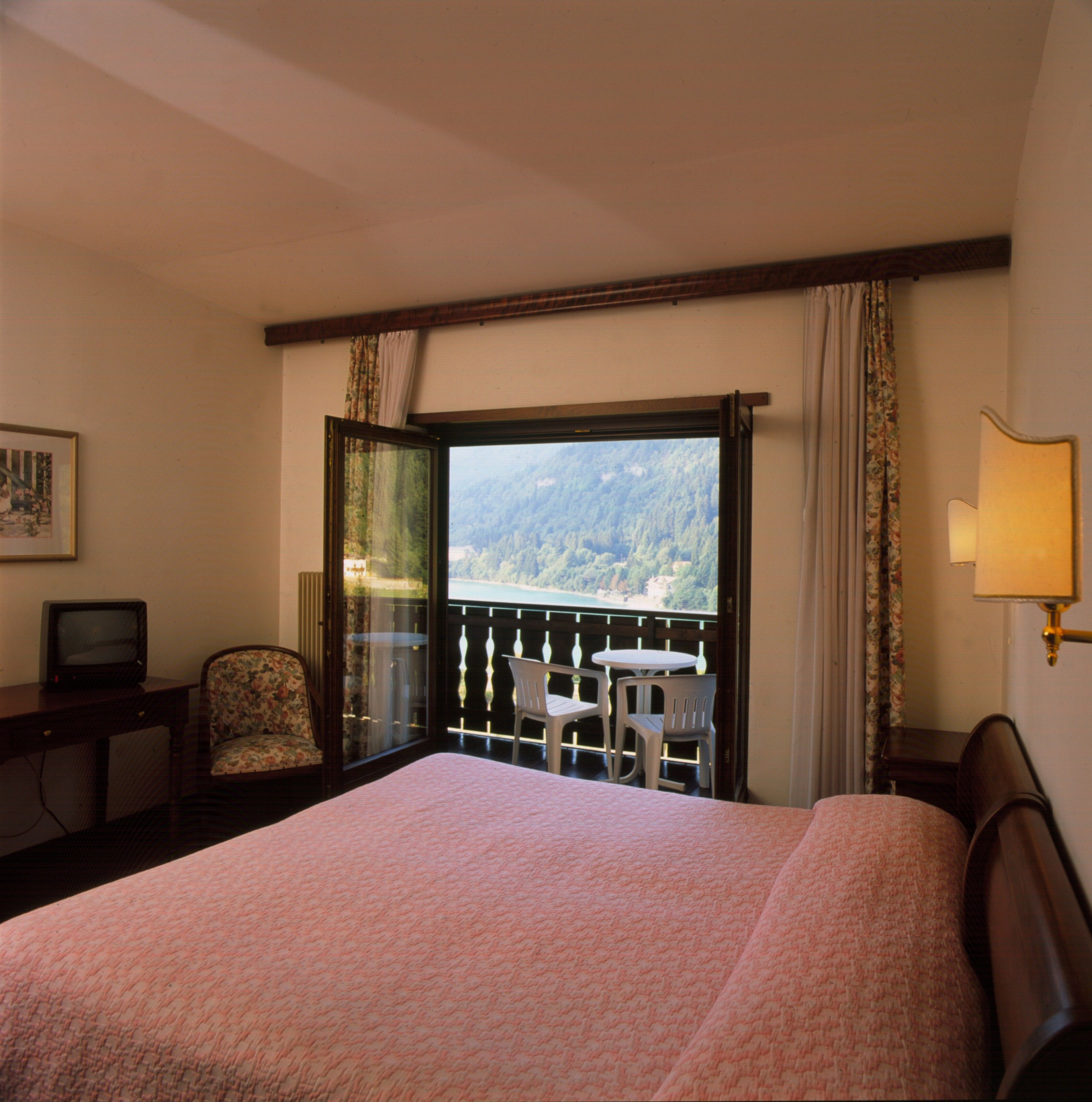 A hotel room with a bed, a desk with a television, a chair, a small table, and a balcony with a view of the mountains.