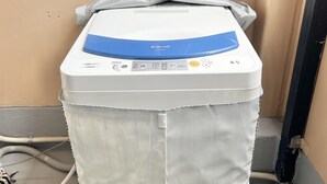 A front-loading washing machine with a control panel, a grey cover, and a hose connected to the side. - Petit Hotel 017 (Tokushima)