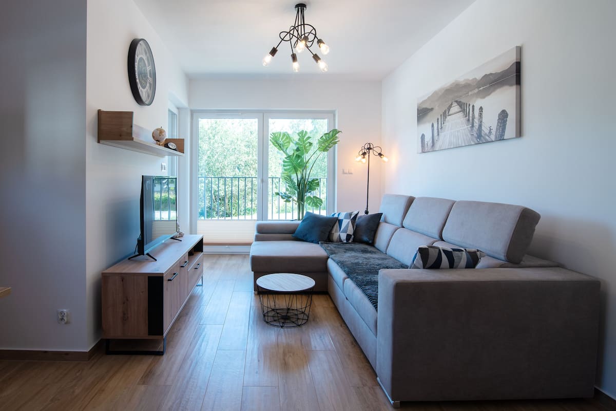 A modern living room with a sofa, a coffee table, a TV, and a large window with a view of greenery.