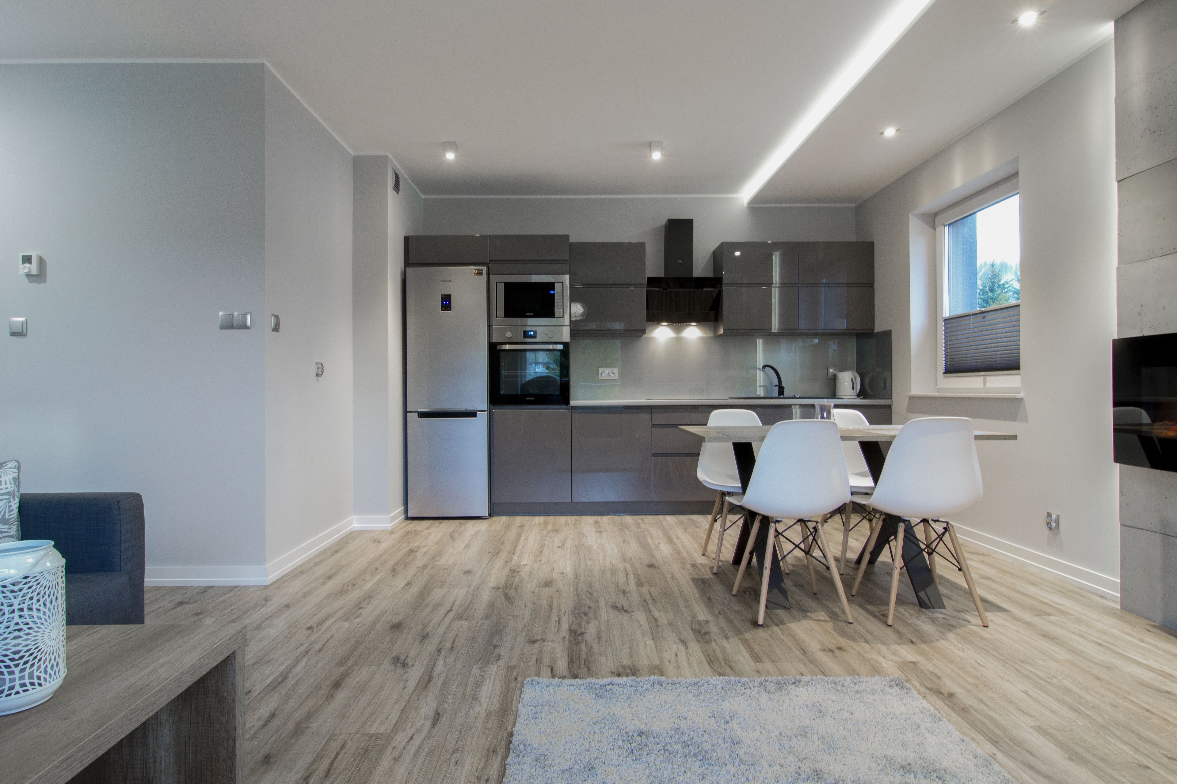 A modern kitchen with stainless steel appliances, a dining area with white chairs, and a wooden floor.