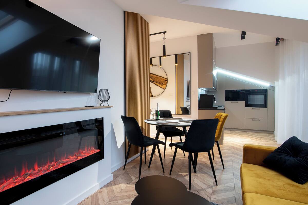 A modern living room with a flat-screen TV, a fireplace, a dining area with chairs, and a kitchen in the background.