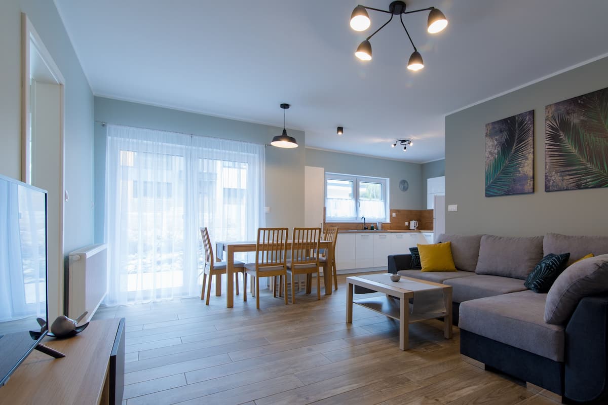 A modern living room with a sofa, coffee table, and chairs. There is a kitchen in the background with a window and a sink.