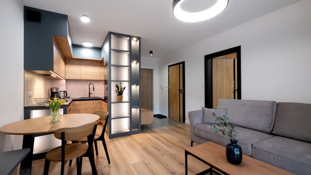A modern living room with a grey sofa, wooden dining table, and chairs. A kitchen with wooden cabinets and a coffee machine is visible in the background. - Apartamenty Sun & Snow Spokojna (Wisla)