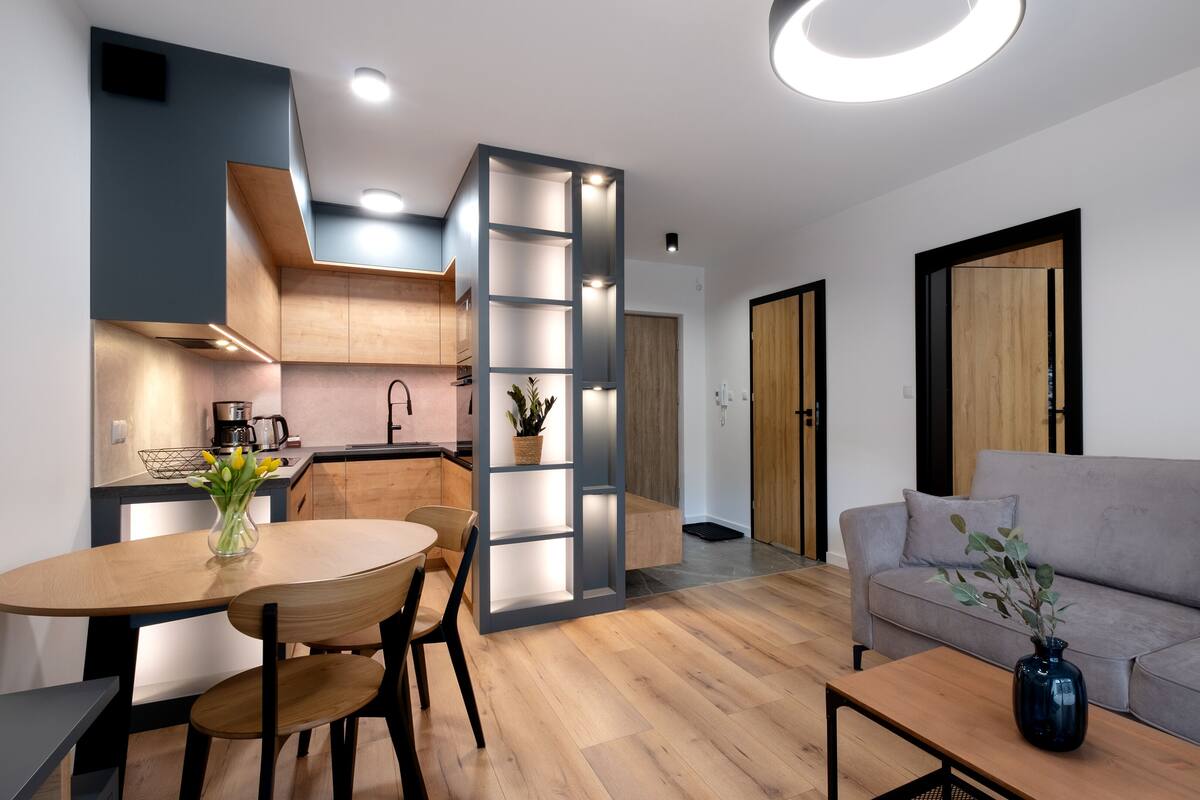 A modern living room with a grey sofa, wooden dining table, and chairs. A kitchen with wooden cabinets and a coffee machine is visible in the background.