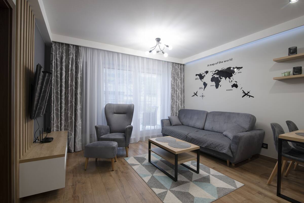 A modern living room with a grey sofa, a grey armchair, a wooden coffee table, and a TV on a wooden stand. There is a world map on the wall and a chandelier.