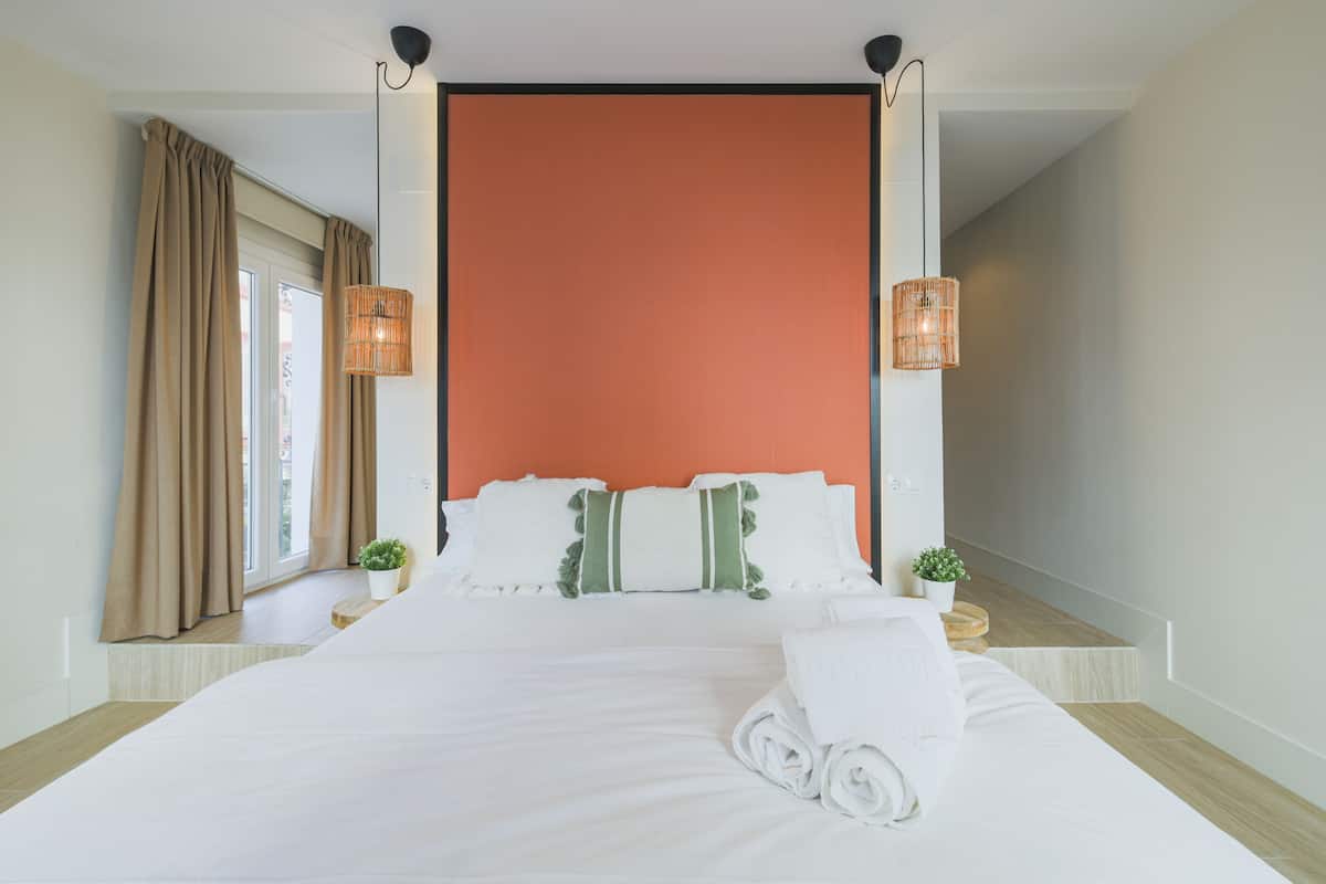A hotel room with a large bed, two bedside tables with potted plants, a window with curtains, and a unique orange accent wall.
