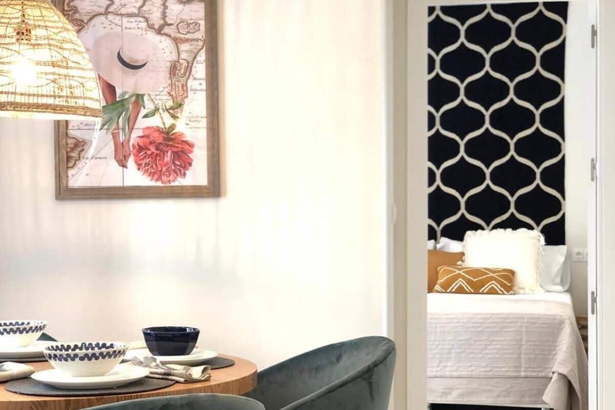 A dining area with a round table, two chairs, and a wall-mounted lamp. A bed with a patterned headboard is visible through an open door.