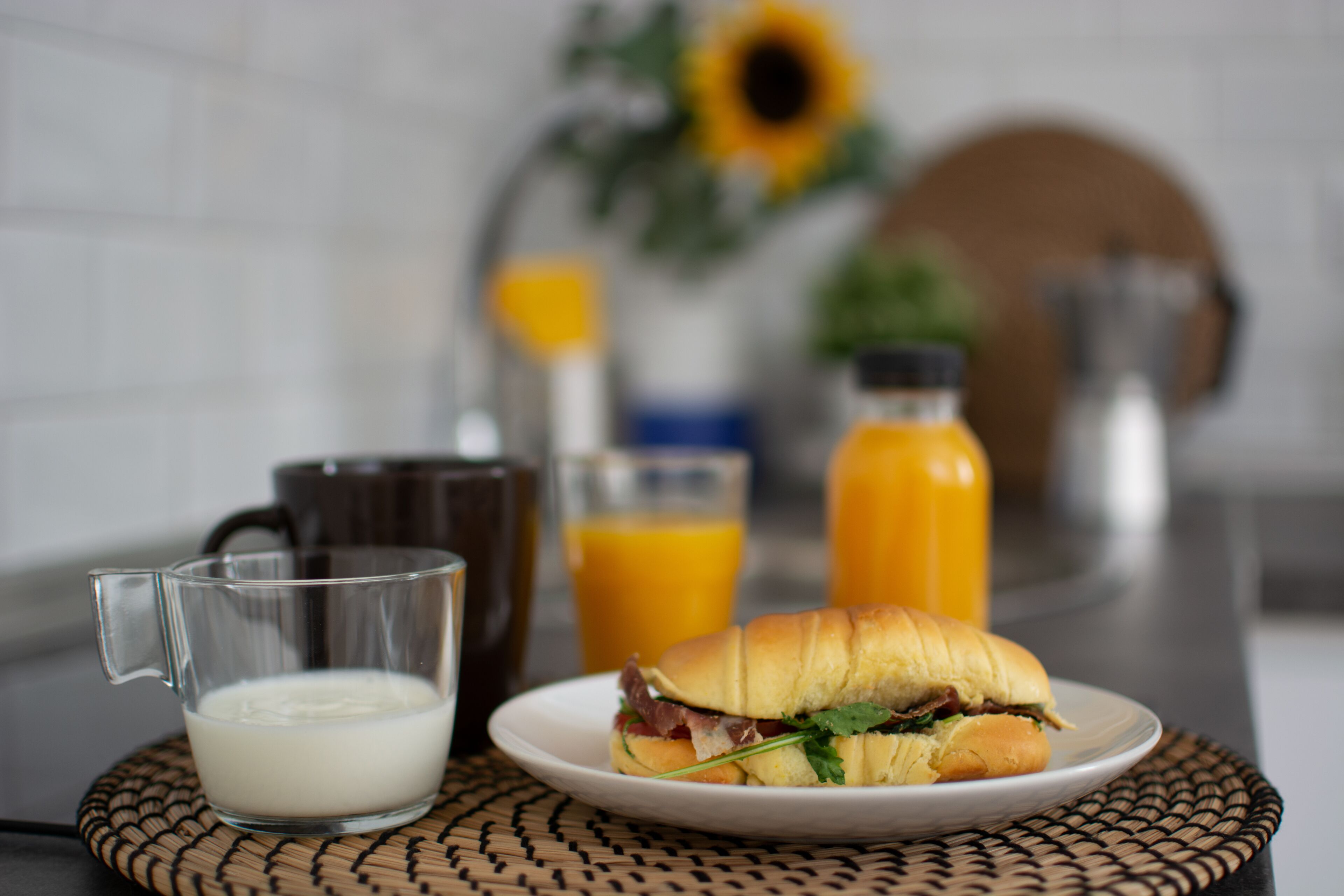 Une table mise pour le petit-déjeuner avec un sandwich, un verre de lait et un verre de jus d’orange, le tout posé sur un set de table en fibres tressées.