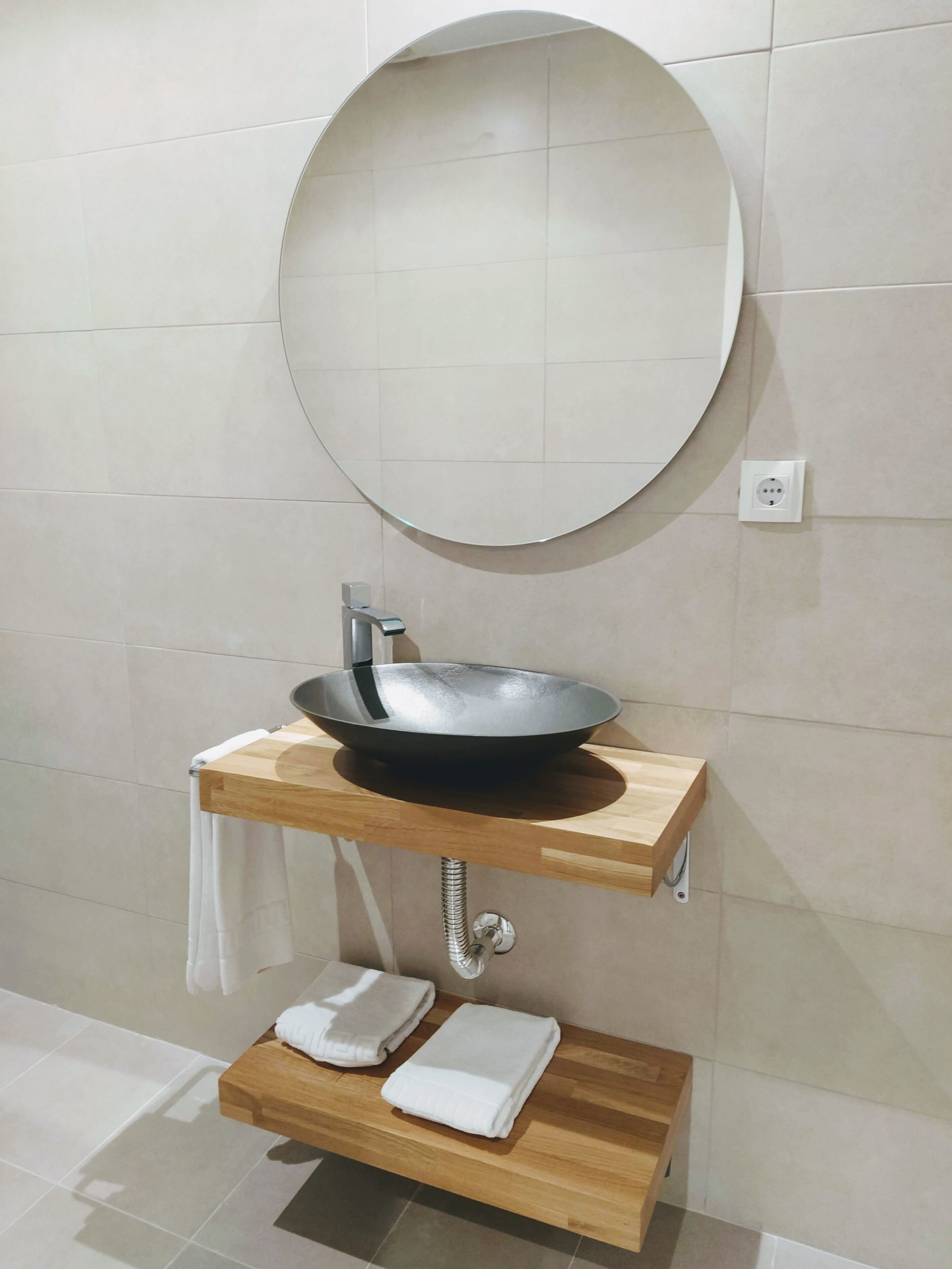 Un bany amb un mirall rodó, un lavabo de forma cilíndrica negre i dues barra de tovalloles de fusta.