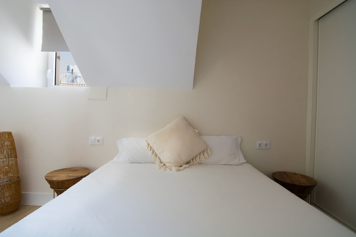 A bedroom with a large bed, two wooden nightstands, a wicker chair, and a view through a window.