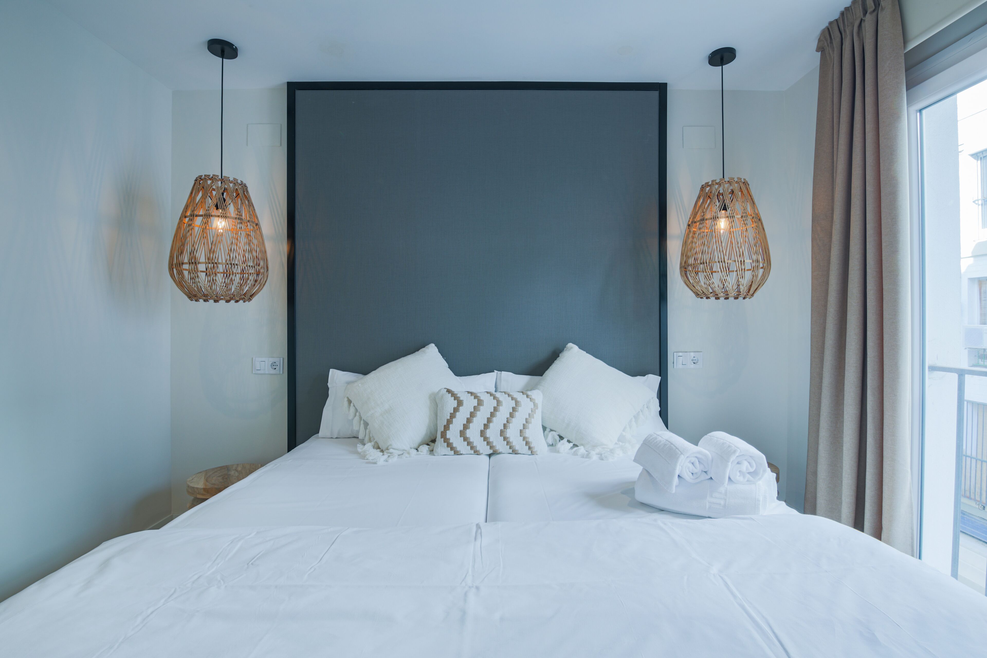 Een moderne hotelkamer met een groot bed, witte lakens, twee hangende rieten lampen en uitzicht buiten door een groot raam.
