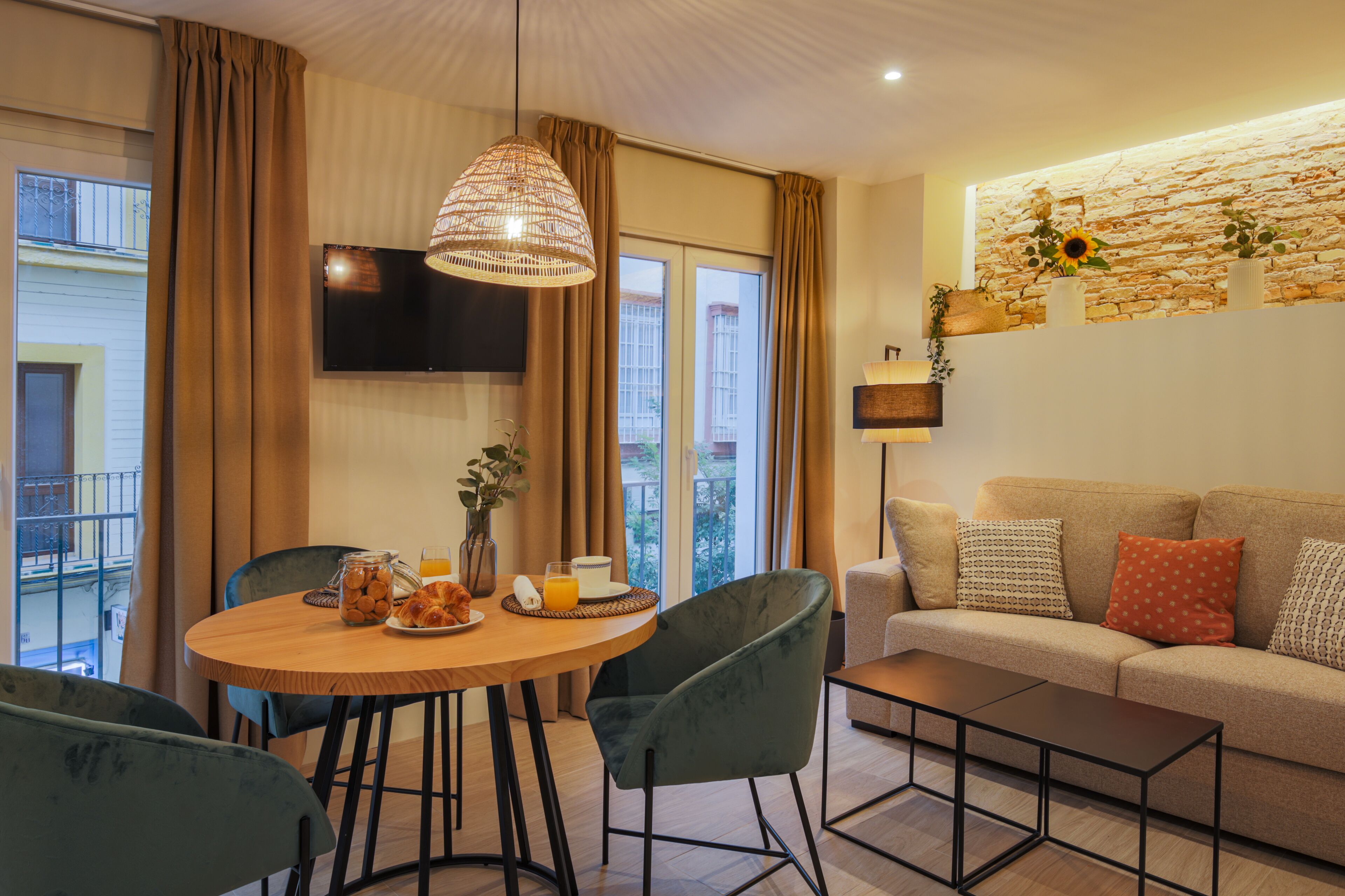 A modern living room with a round wooden table, green chairs, a sofa with patterned cushions, a mounted TV, and a stone wall with plants.