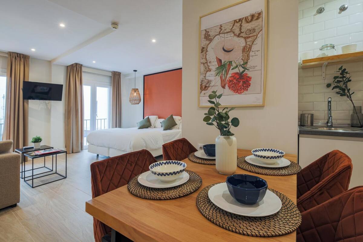 A modern hotel room with a bed, a TV, a dining area with a table set for two, and a kitchen area.