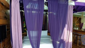Una habitación con una cama, cortinas moradas, suelo de madera, un televisor y una pequeña mesa con un jarrón de flores.