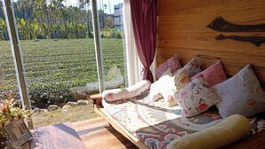 Una cama de madera con ropa de cama floral, una mesa de madera con una revista y un control remoto, y vistas a un campo verde a través de amplias ventanas.
