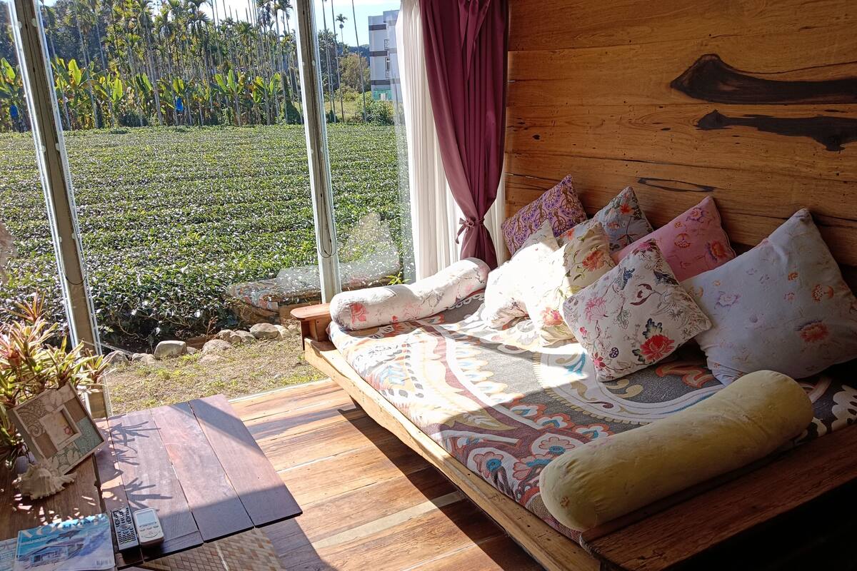 A wooden bed with floral bedding, a wooden table with a magazine and remote control, and a view of a green field through large windows.