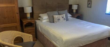 Un dormitorio con una cama grande, dos mesitas de noche con lámparas, una silla de mimbre y cuadros enmarcados en la pared.
