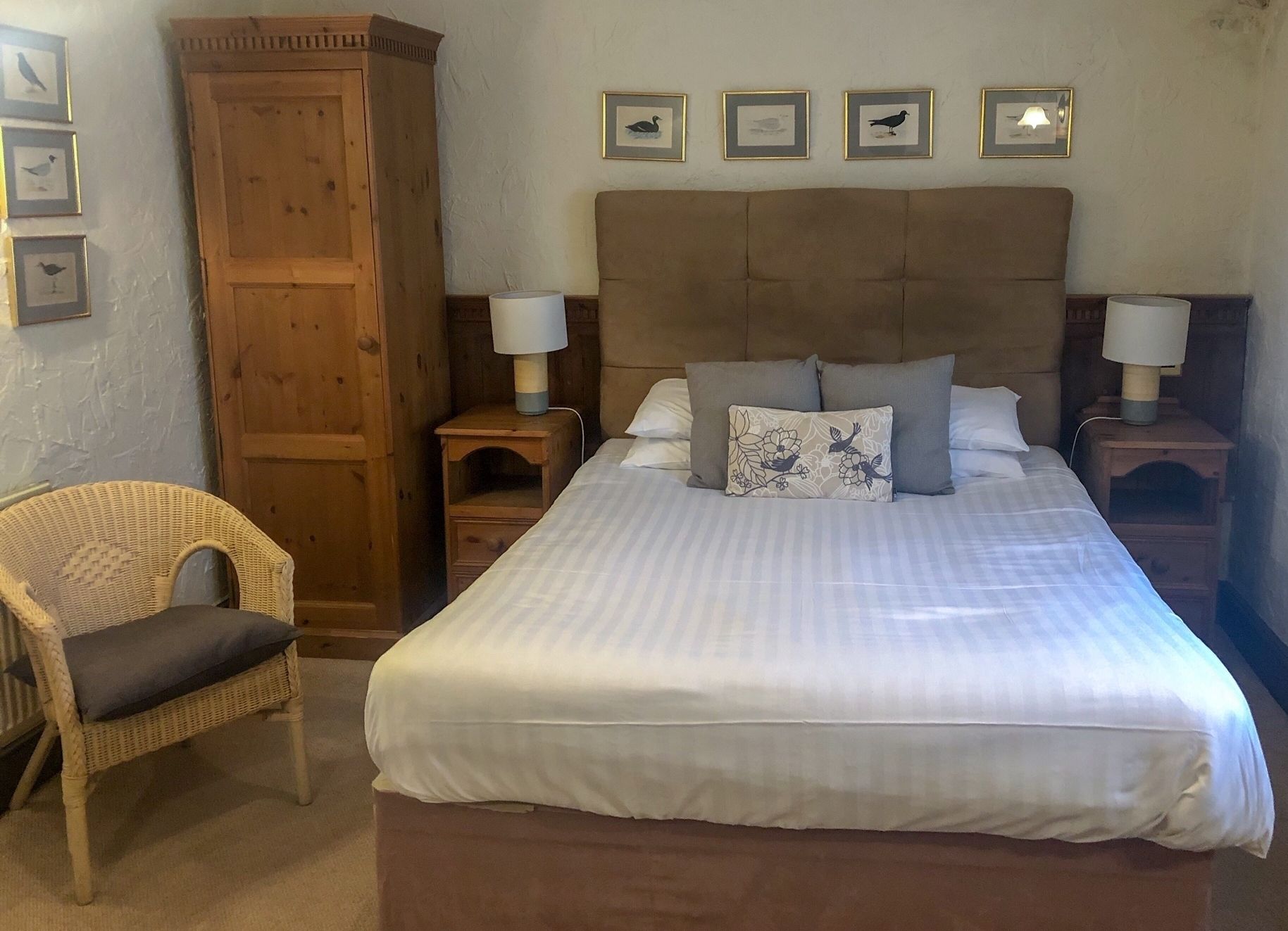 A bedroom with a large bed, two bedside tables with lamps, a wooden wardrobe, a wicker chair, and framed pictures on the wall.