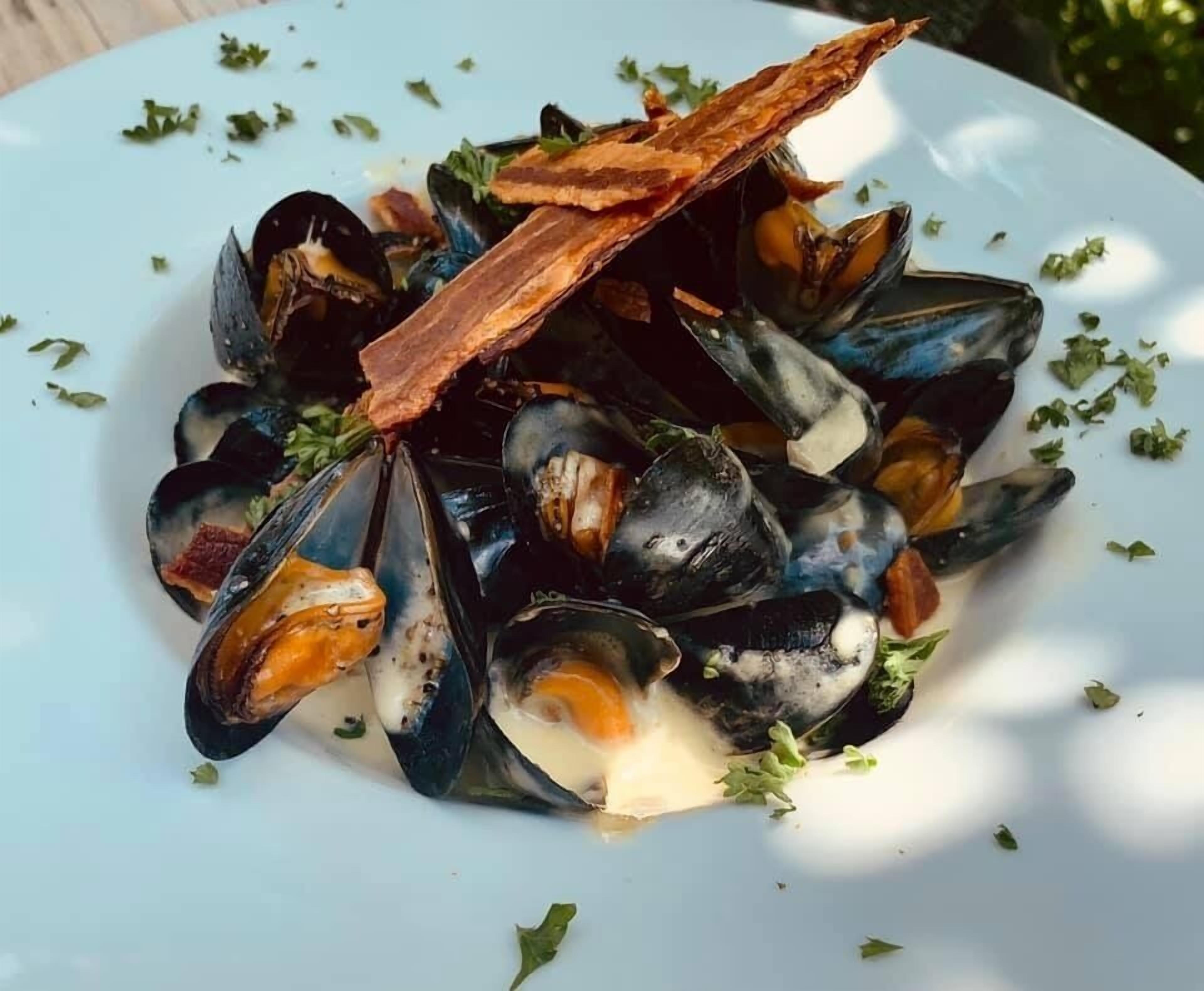 A plate of mussels with a piece of bacon on top, garnished with herbs.