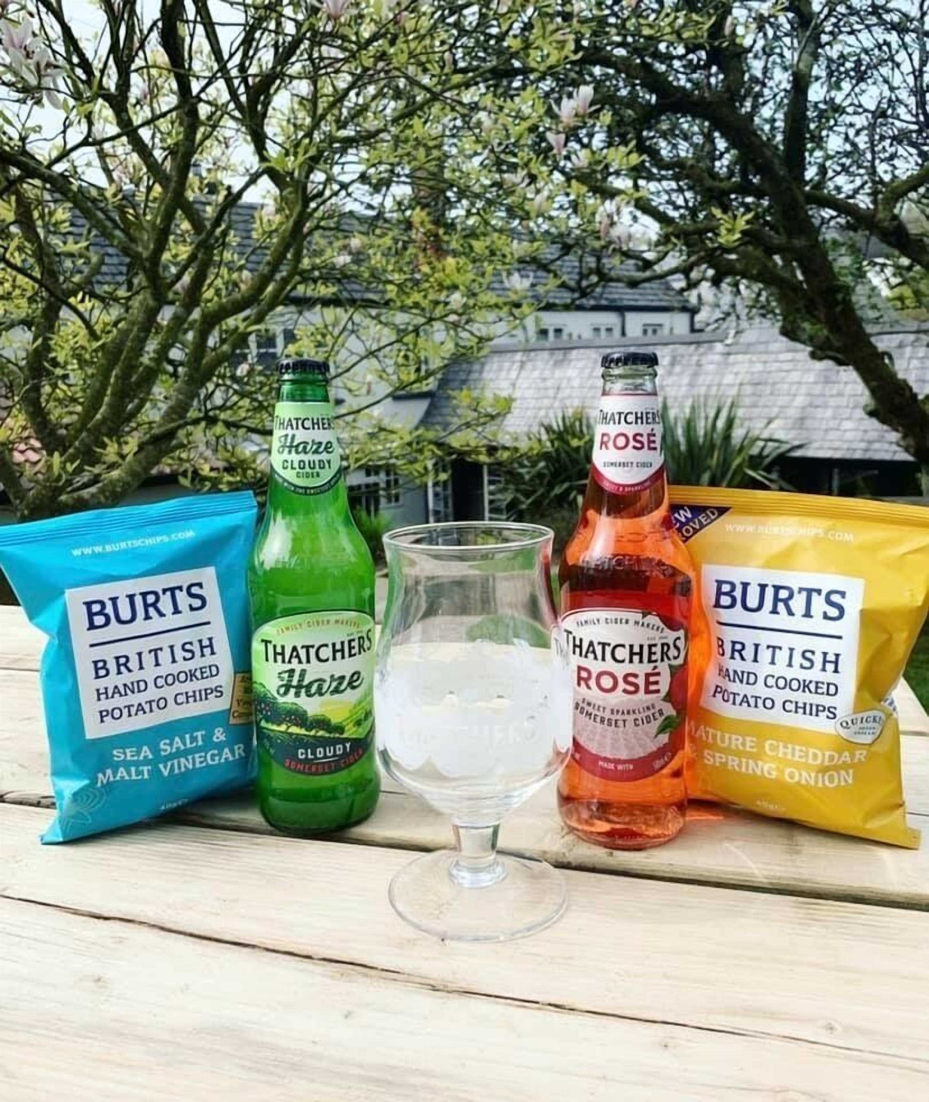 A table with two bags of Burts British hand cooked potato chips, two bottles of Thatchers Haze Cloudy and Thatchers Rose sparkling cider, and a glass of ice.
