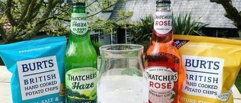 A table with two bags of Burts British hand cooked potato chips, two bottles of Thatchers Haze Cloudy and Thatchers Rose sparkling cider, and a glass of ice.
