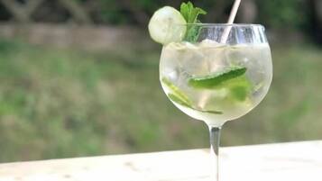 Un vaso transparente con pajita, conteniendo una bebida con hielo y hojas de menta, sobre una superficie de madera con un fondo borroso de vegetación.
