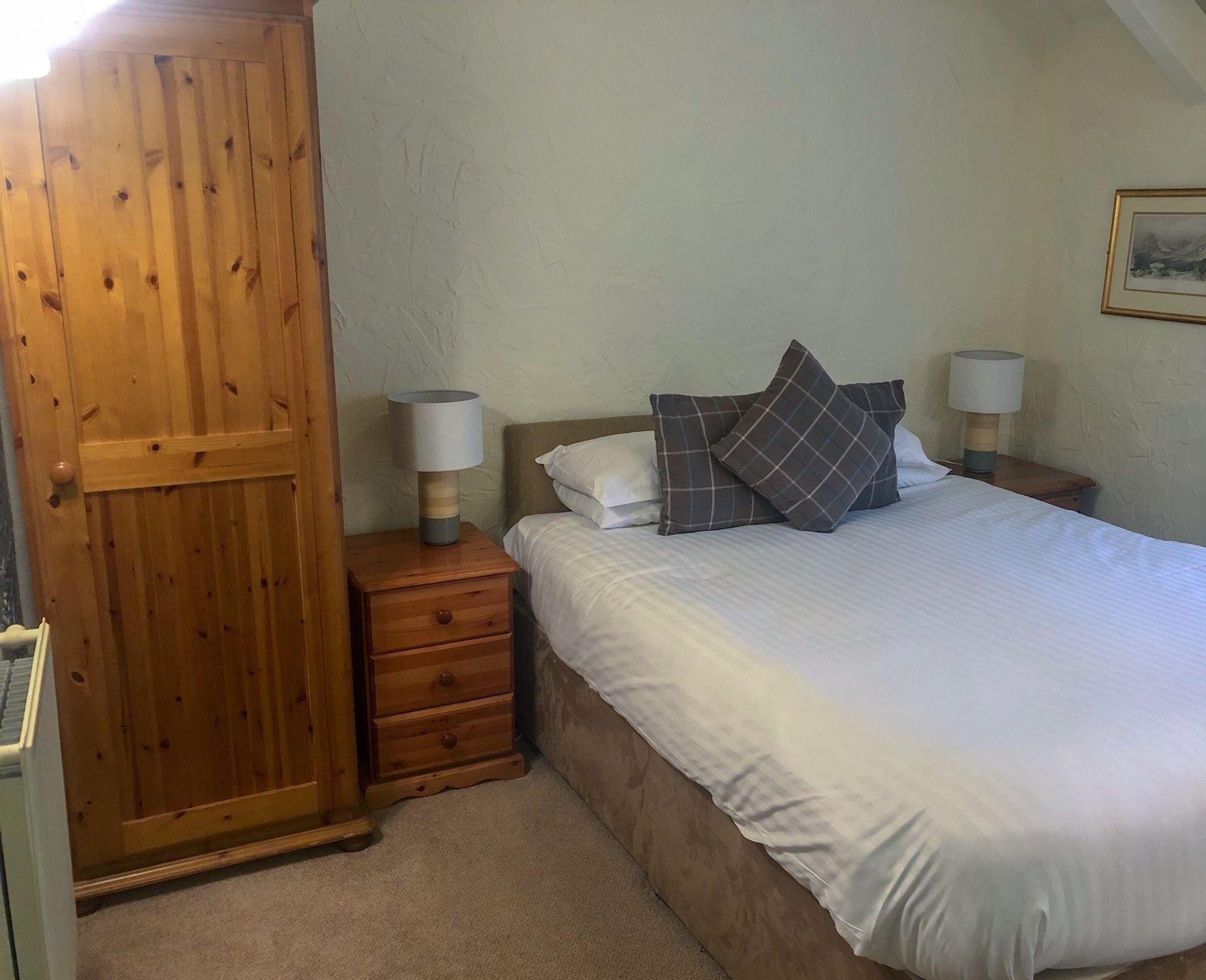 A bedroom with a wooden wardrobe, a bed with white bedding and plaid pillows, two bedside lamps, and a framed picture on the wall.