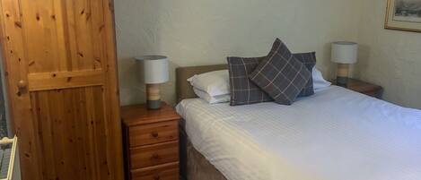 A bedroom with a wooden wardrobe, a bed with white bedding and plaid pillows, two bedside lamps, and a framed picture on the wall.