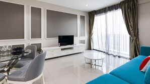 A modern living room with a glass dining table, grey upholstered chairs, a white entertainment unit with a flat-screen TV, a blue sofa, and a large window with sheer curtains. - Luxury Suite Icity Shah Alam (Shah Alam)