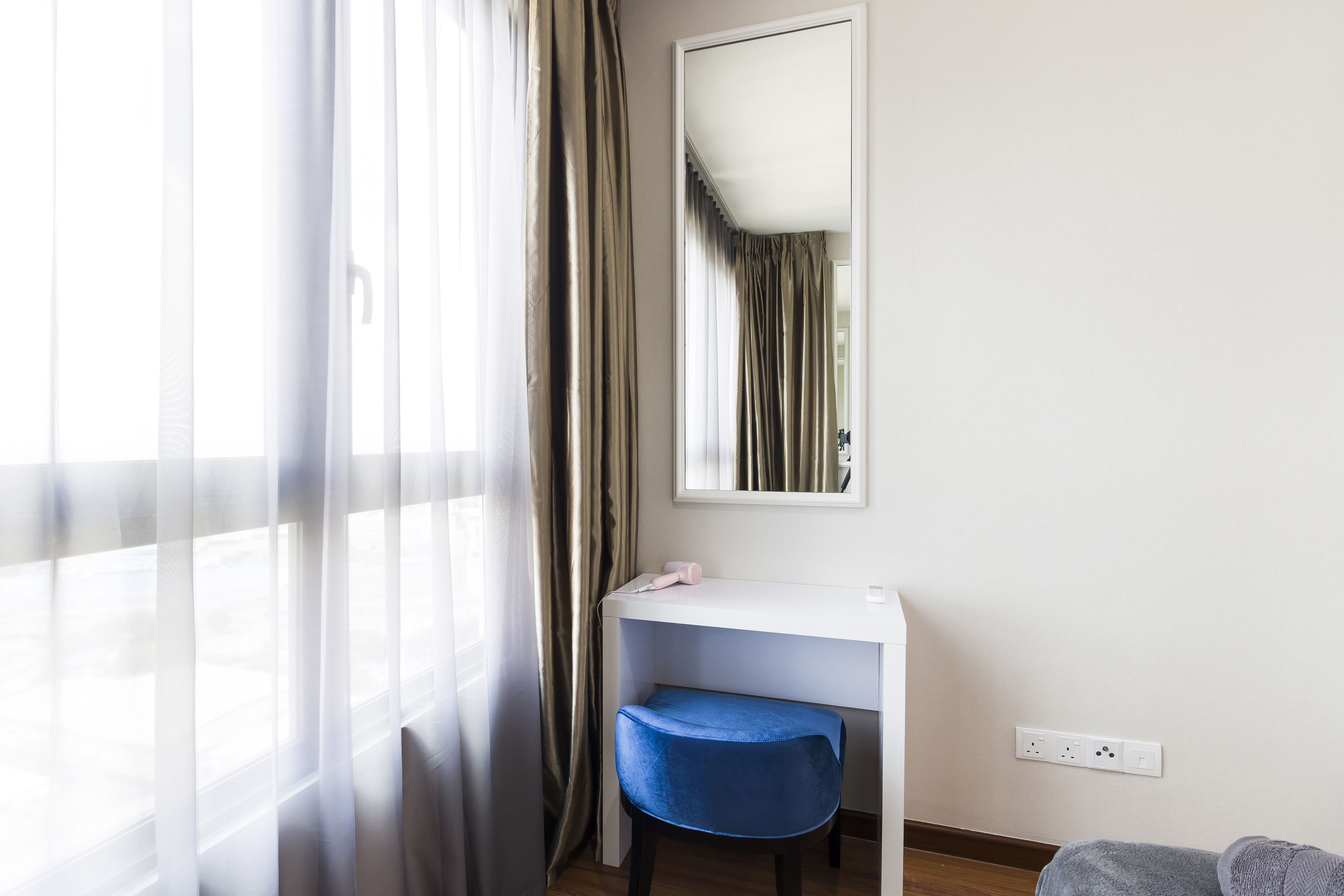 A hotel room with a white desk, a blue chair, a mirror, and a window with sheer curtains.