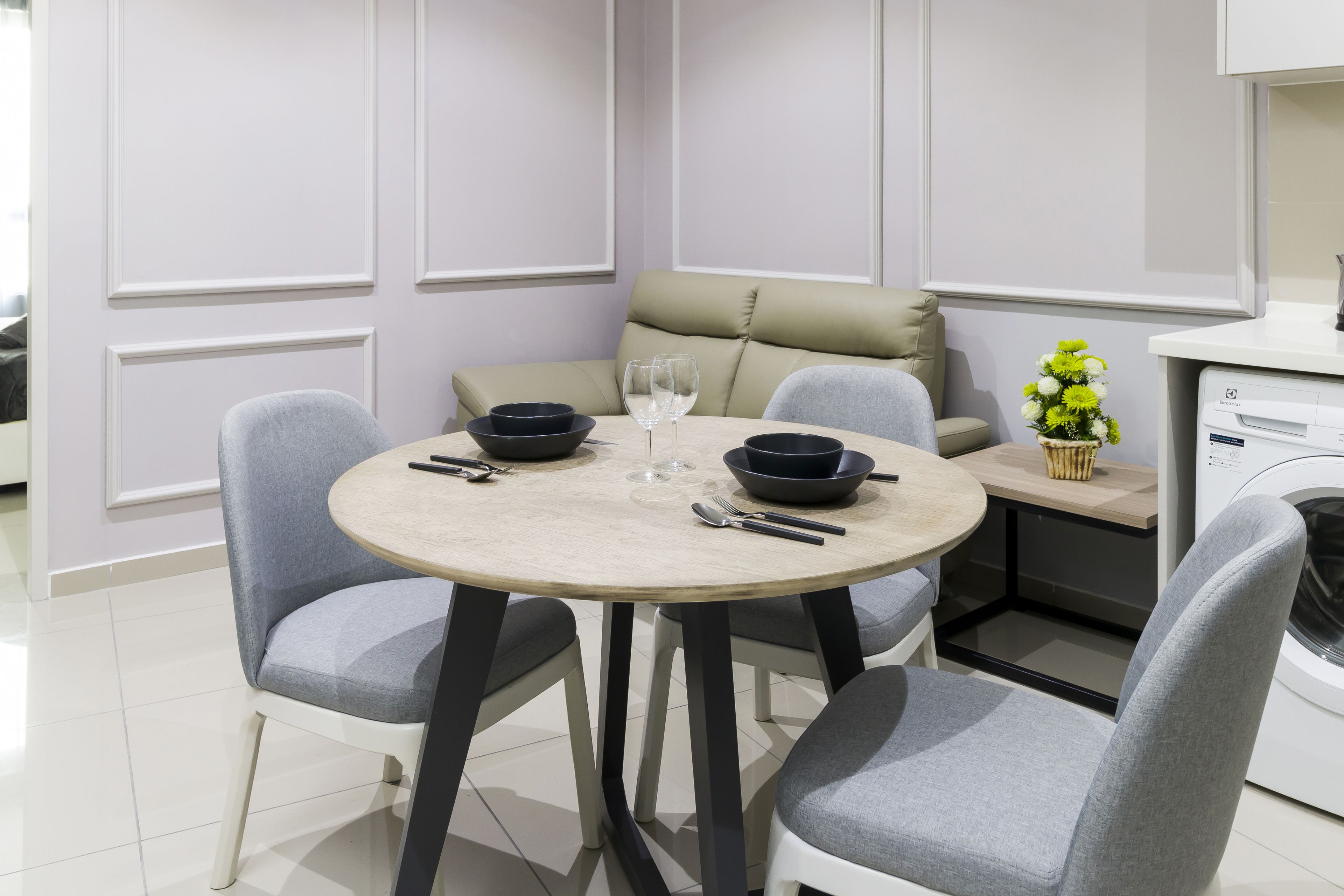 A modern dining area with a round table, grey chairs, a beige sofa, and a small side table with a vase of flowers.
