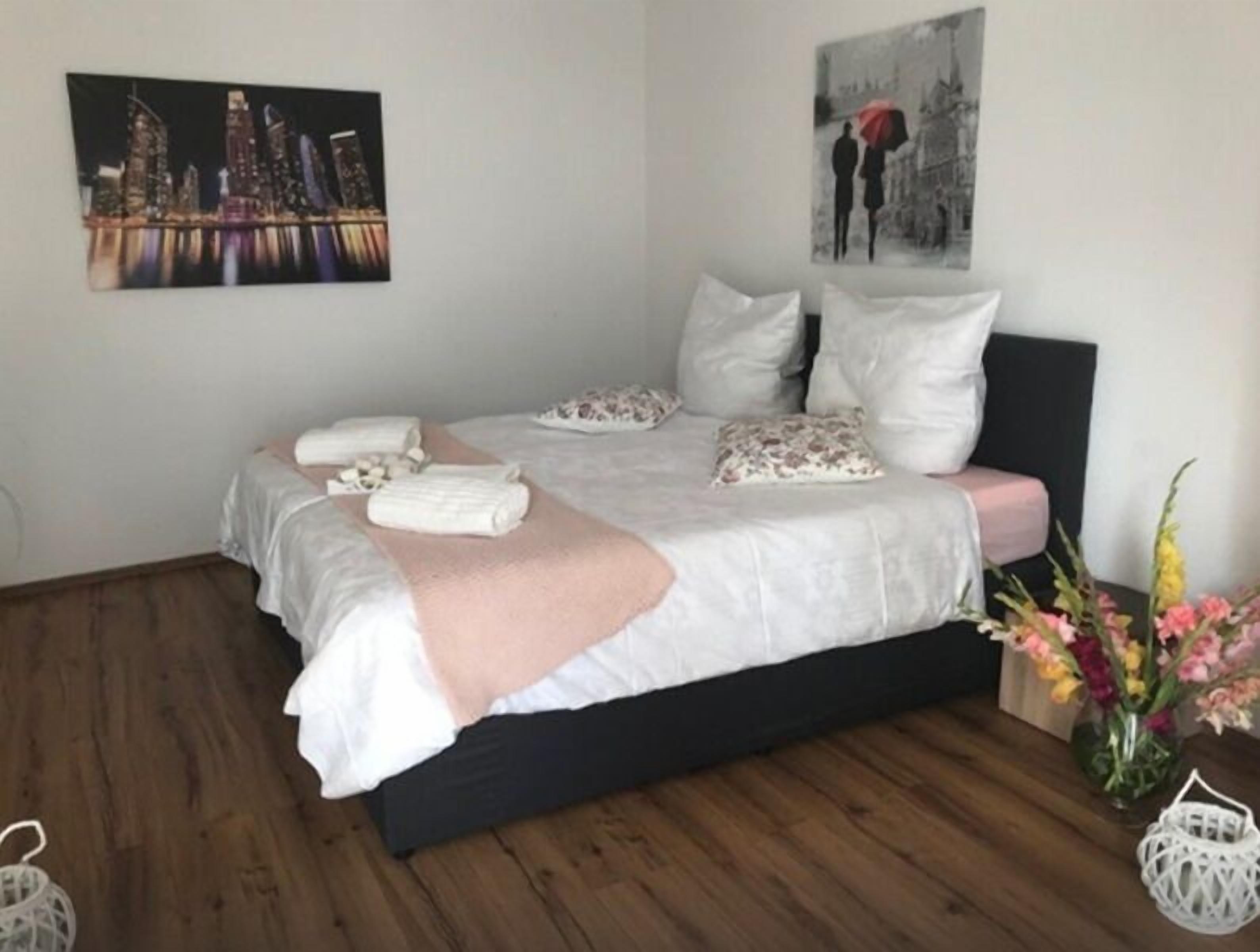 A neatly made bed with white linens and a pink throw, a cityscape painting on the wall, and a floral arrangement on the side.