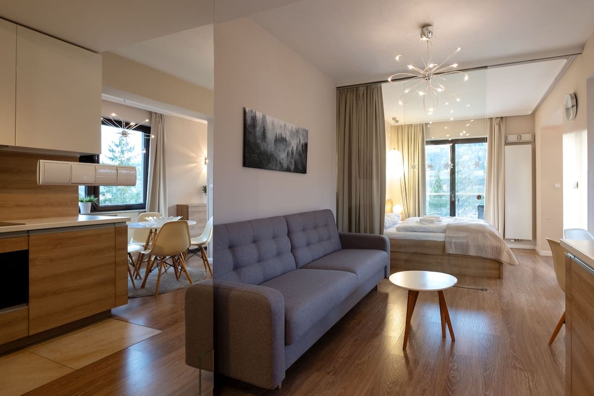 Una habitación de hotel moderna con sofá, una pequeña mesa redonda, cama con vistas, zona de cocina y comedor.