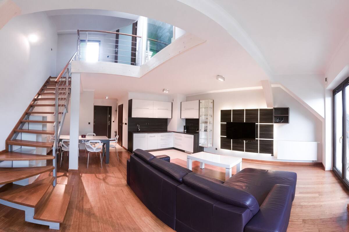 Un salón comedor amplio con una escalera que sube a un nivel superior, una cocina con electrodomésticos modernos y un televisor grande montado en la pared.