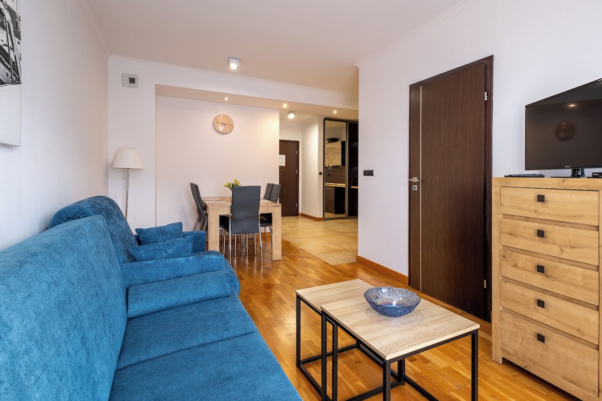 Una sala moderna con un sofá azul, un mueble de madera con cajones, un televisor y un comedor con mesa y sillas.