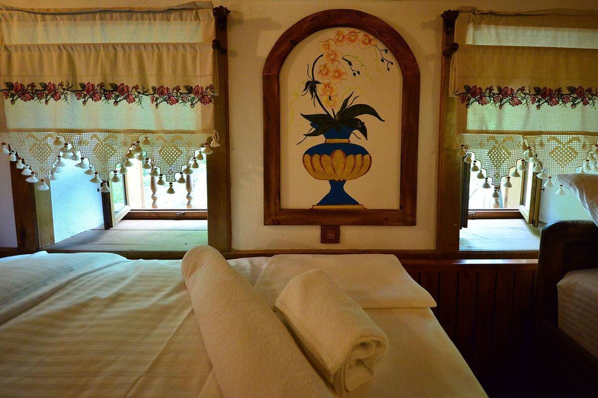 Une chambre d’hôtel avec deux lits, une grande fenêtre ornée de rideaux en dentelle, un tableau floral encadré et un luminaire suspendu.