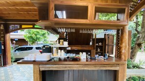A wooden outdoor bar area with a patterned floor, shelves with glassware, and a view of a parked car and greenery in the background.