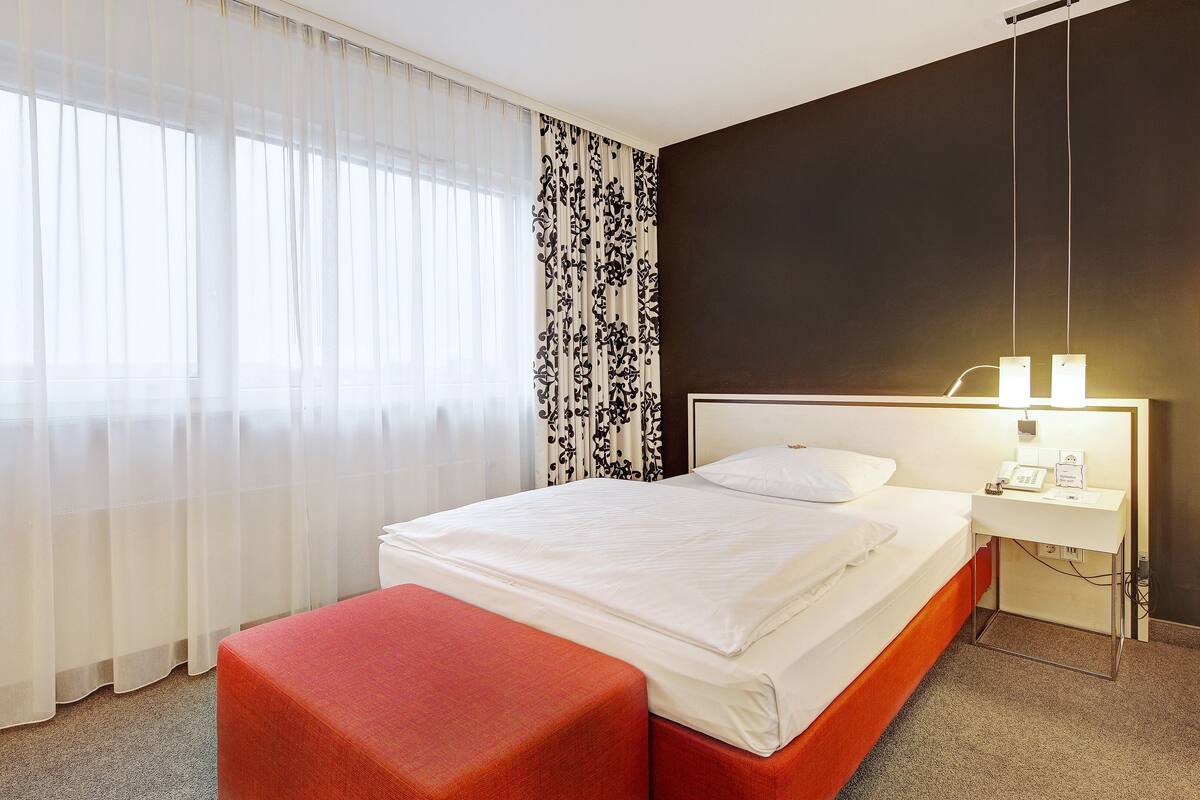 Une chambre d’hôtel avec un lit, une table de chevet avec une lampe, un pouf rouge et des rideaux transparents.