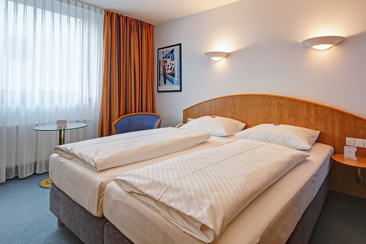 Une chambre d’hôtel avec deux lits, une chaise, une petite table et une table de chevet fixée au mur.