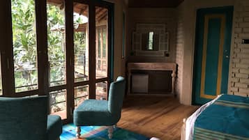 Quarto com piso de madeira, cama com colcha listrada, uma cadeira, uma pequena mesa e janela com vista para o verde.