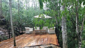 Um deck de madeira com cadeiras e uma mesa brancas, rodeado por vegetação exuberante e um pequeno lago.