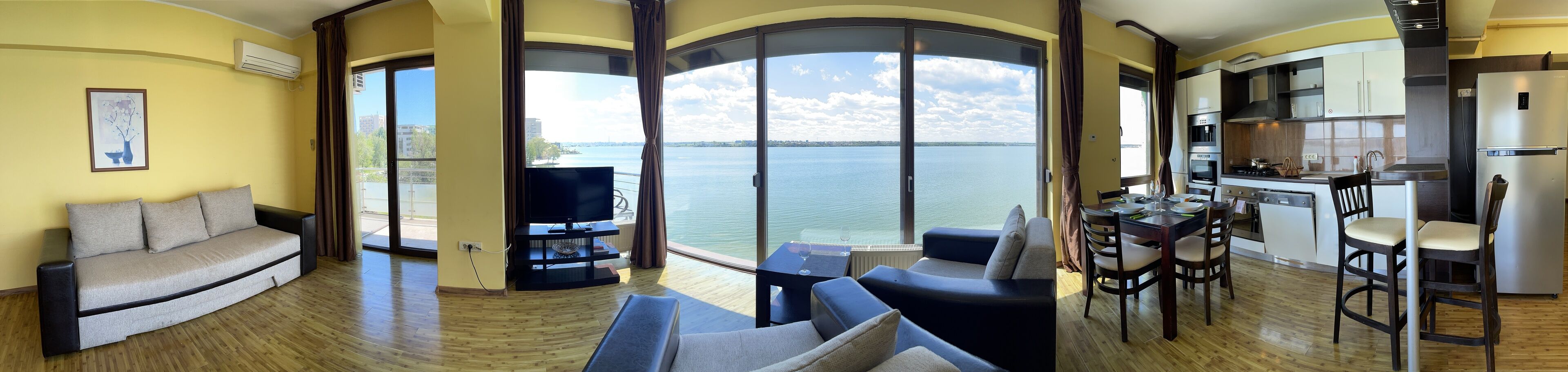 Un vaste espace de vie comprenant un canapé, un téléviseur et une table à manger avec des chaises. Une grande fenêtre offre une vue sur l’eau.