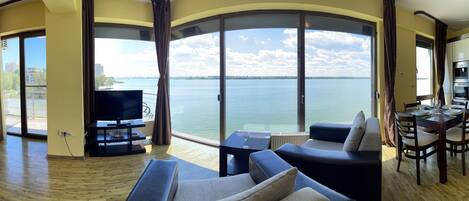 A spacious living area with a sofa, a TV, and a dining table with chairs. A large window offers a view of the water.