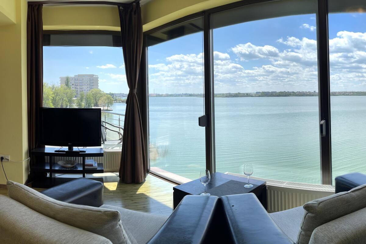 Una sala de estar moderna con un sofá, un televisor y una mesa con una copa de vino. Un ventanal grande ofrece vistas a una masa de agua y edificios.