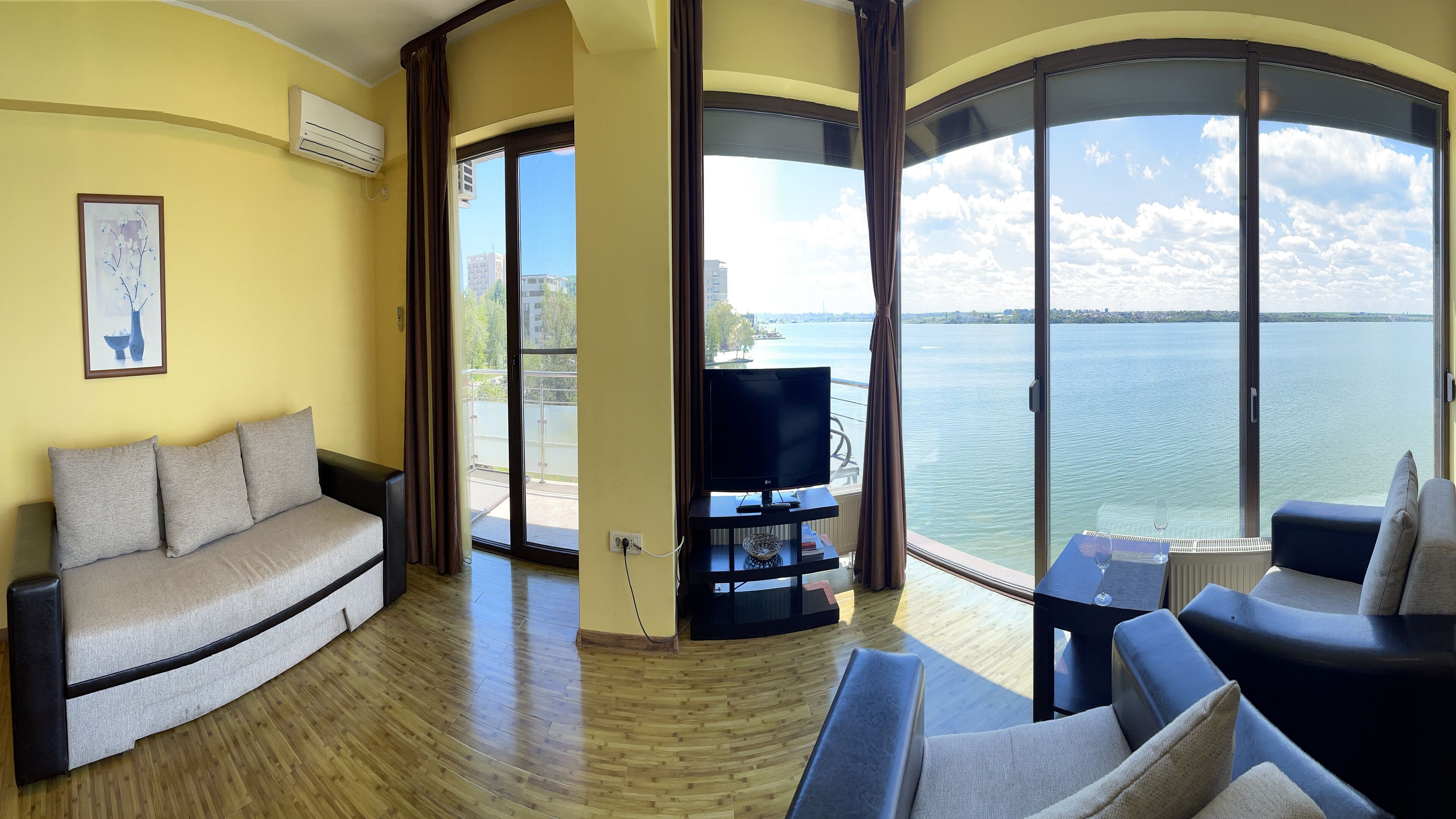 A living room with a sofa, a TV on a stand, and a view of a body of water through large windows.