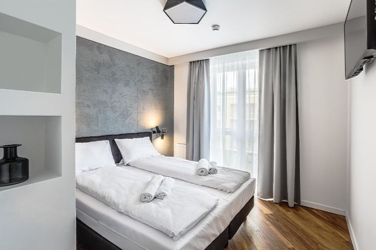 A modern hotel room with a large bed, a grey accent wall, a flat-screen TV, and a large window with curtains.