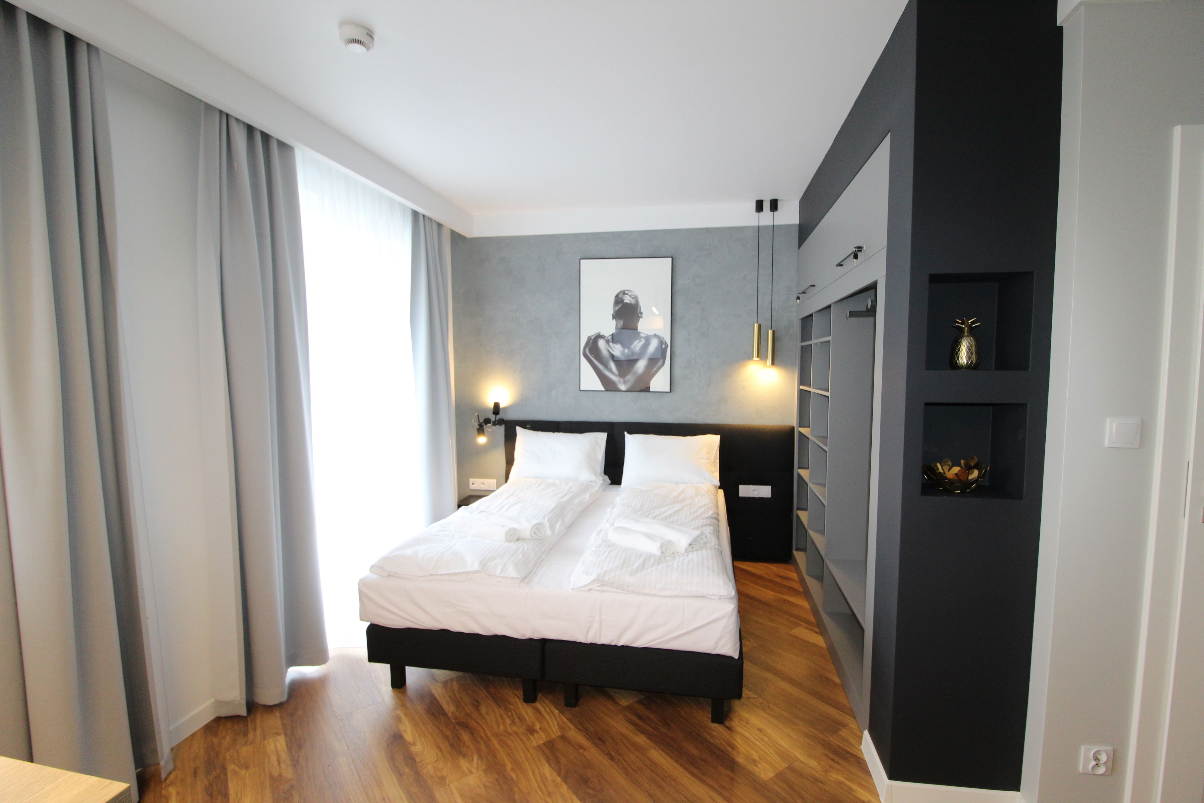 A modern bedroom with a large bed, grey curtains, a wall-mounted light fixture, and a small shelf with decorative items.