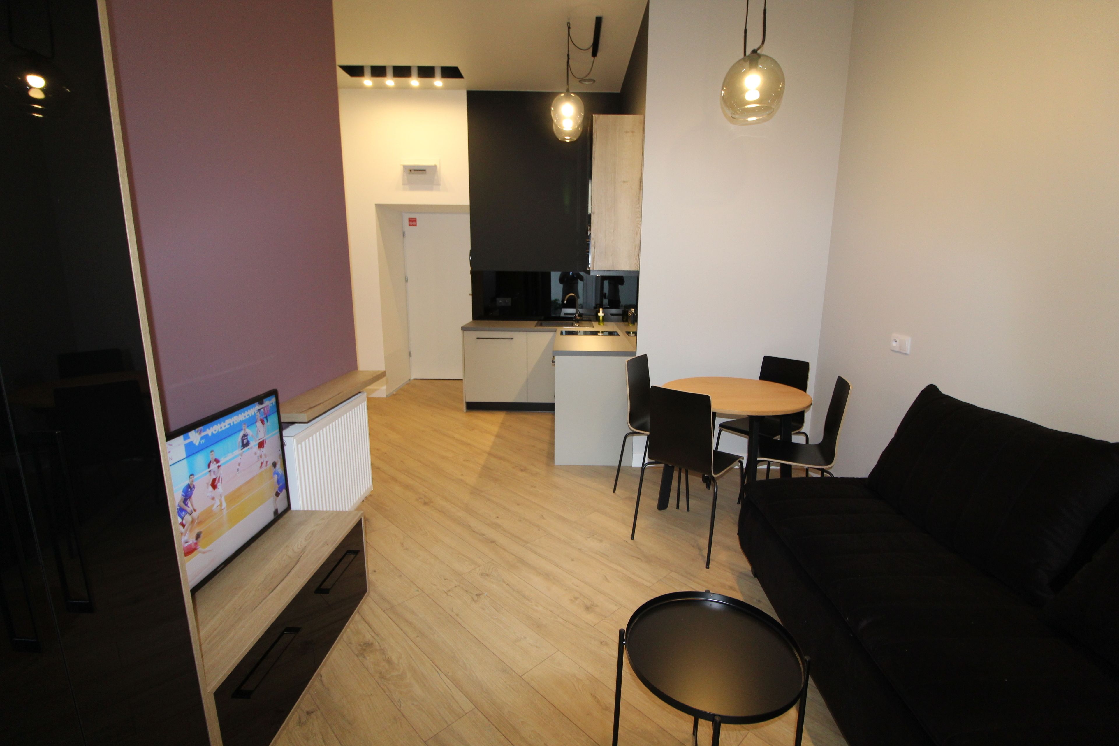 Un moderno salón con un sofá negro, una mesa de centro de madera y un televisor que muestra un partido deportivo. Se ve una cocina con zona de comedor al fondo.
