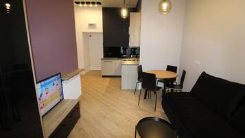 Un moderno salón con un sofá negro, una mesa de centro de madera y un televisor que muestra un partido deportivo. Se ve una cocina con zona de comedor al fondo.