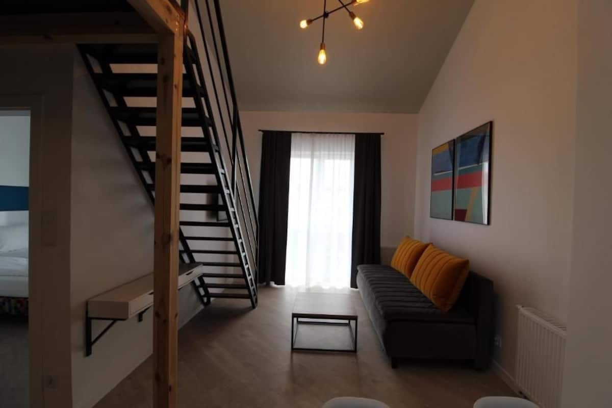 A modern living room with a staircase, a sofa with orange cushions, a coffee table, and a painting on the wall.
